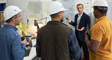 Middle aged Caucasian man presenting building project plan on whiteboard to group of young adult multiethnic men wearing safety helmets in office setting, discussing construction details